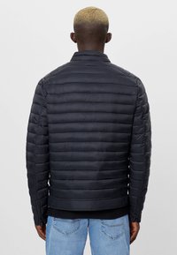 Man med kort blont hår som bär en svart kviltad jacka och ljusblå jeans, sedd bakifrån mot en enfärgad bakgrund.