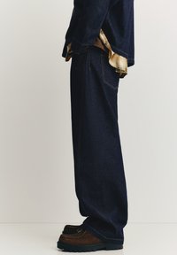 Temno modre kavbojke iz jeansa z ravnim izrezom noge, z vidnimi šivi, kombinirane z rjavimi čevlji in karirasto srajco spodaj.