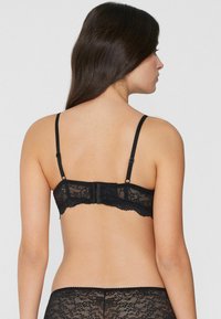 Černá krajková podprsenka s nastavitelnými ramínky, vroubkovaným lemováním a zapínáním na háček a očko. Krajkový vzor je výrazný a ukazuje složité detaily.