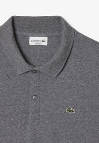 Grå polo skjorta med ribbad krage, treknappsknäppning och liten grön krokodil-logga på vänster bröst. Tillverkad av textil med struktur.