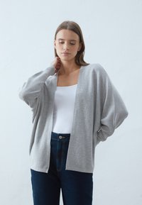 Světle šedý kardigan vyrobený z pletené látky, oversize design s otevřeným předním dílem, dlouhými rukávy a ribbanou texturou. Nosený přes bílé tričko a džíny.
