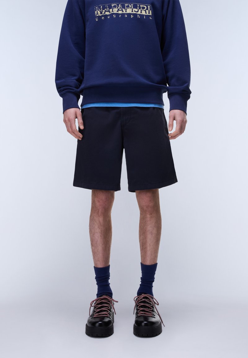 Άνδρας που φοράει μπλε σκούρο φούτερ με τυπωμένο λογότυπο, μαύρο σορτς, σκούρες μπλε κάλτσες και μαύρα παπούτσια με κορδόνια, στέκεται σε απλό φόντο.