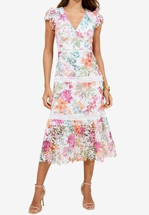 Femme portant une robe midi blanche sans manches avec un imprimé floral multicolore et des détails en dentelle, assortie à des talons nude à bride à la cheville et un bracelet en or.