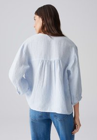 Lichtblauwe, loszittende blouse van textuurstof met driekwartmouwen en een geplooide achternaad. Draagt het met blauwe jeans.