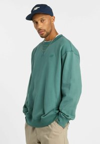 Sweatshirt vert à col rond et poignets côtelés, avec un logo texturé sur la poitrine. Porté avec une casquette marine et un collier en or.