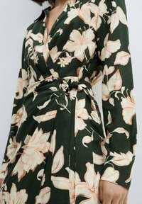 Zelena haljina na preklop s dugim rukavima, s cvjetnim uzorkom u mekim ružičastim tonovima, detaljem s vezivanjem u struku i nabranim dijelom suknje.