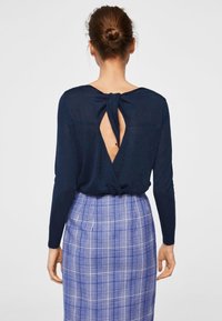 Kobieta z włosami w kok, ubrana w granatową bluzkę z długim rękawem i węzłem z tyłu oraz w niebieską szkocką spódnicę, stojąca na jednolitym tle.