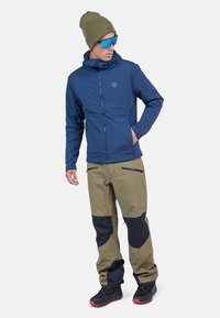 Marineblaues Jacket mit Reißverschlüssen, leichtem Material und Kapuze; khakifarbene Hose mit schwarzen Akzenten, mehreren Taschen und Sneakers.