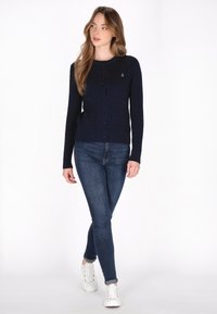 Marineblauwe ribgebreide cardigan met een ronde hals, knoopsluiting en een klein ankerembleem op de borst, gecombineerd met skinny jeans en witte sneakers.