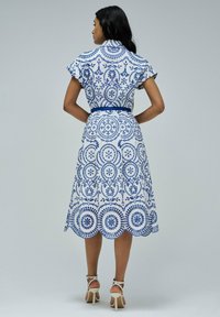 Vestido blanco con intrincados patrones florales azules, mangas cortas y una cintura ceñida con cinturón. El dobladillo presenta bordes en forma de conchas. Se lleva con tacones blancos y con tiras.