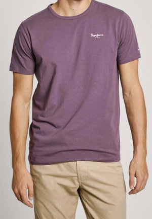 Homme portant un t-shirt violet uni à manches courtes avec un petit logo blanc sur la poitrine et un pantalon beige, debout les bras détendus le long du corps.