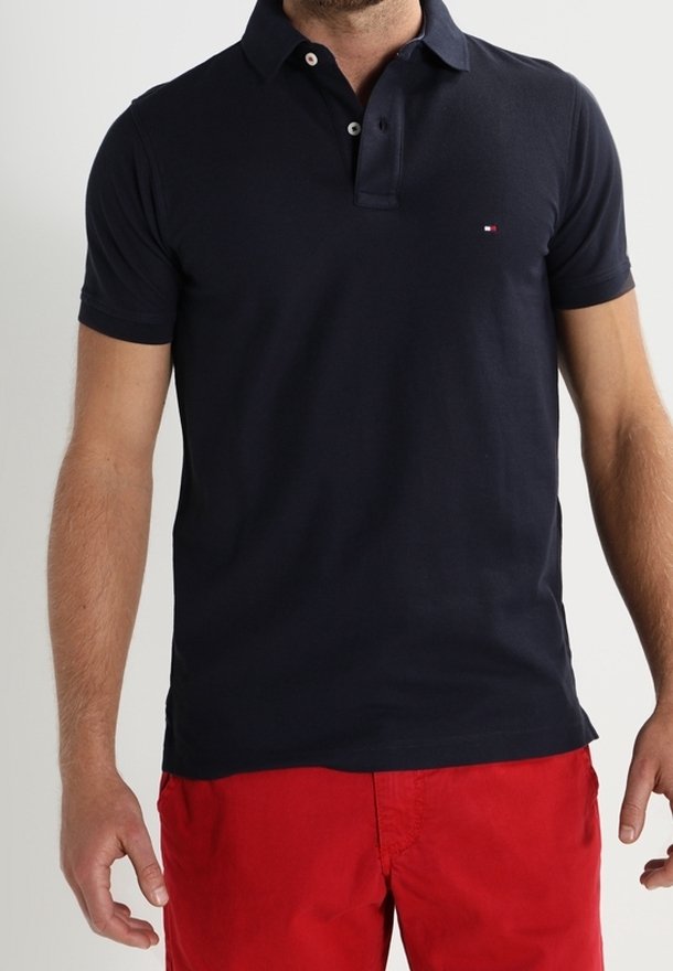 Polo shirt bleu marine en coton, avec un col à deux boutons, des manches courtes et un petit logo sur la poitrine. Assorti avec un short rouge.