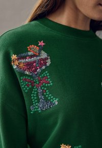 Sweatshirt verde cu un design texturat, brodat, al unui pahar de cocktail, având paiete, flori și un accent în formă de stea.