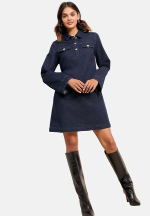 Femme portant une robe en denim sombre avec des poches poitrine boutonnées et des manches évasées, associée à des bottes en cuir noires montantes.