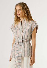 Ženska z valovitimi lasmi, oblečena v brezrokavnik s pasom in črtastimi vzorci ter cvetličnim čipkastim detajlom, oblečene bele hlače, ki rahlo gleda v stran.