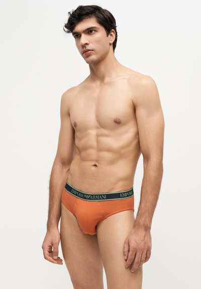 Jeune homme aux cheveux foncés portant un slip orange Emporio Armani, debout devant un fond uni clair, regardant légèrement sur le côté.