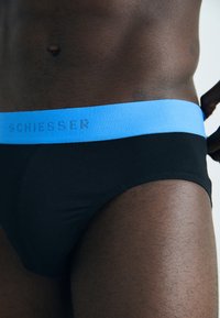 Uomo che indossa slip neri con una larga fascia blu etichettata "SCHIESSER," mentre aggiusta la fascia.