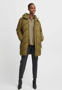 Cappotto imbottito verde oliva con cappuccio, caratterizzato da un design trapuntato, chiusura con zip e tasche laterali, indossato sopra una camicia a righe e pantaloni scuri.