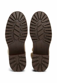 Suela de goma marrón con patrón de zigzag y surcos profundos para tracción. Presenta una parte superior de ante en color beige neutro y un forro suave.