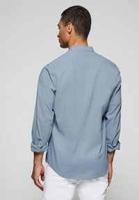 Ljusblå långärmad skjorta med rak nederkant och rullade manschetter, gjord av ett mjukt tyg; bakifrån syns en klassisk krage.