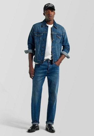 Giovane uomo che indossa una giacca di jeans blu scuro e jeans, t-shirt bianca, scarpe nere lucide e un berretto nero con la scritta "Milano 1984".