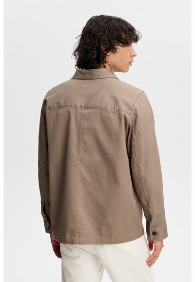 Mężczyzna z kręconymi włosami, ubrany w beżową, długorękawową kurtkę i białe spodnie, stojący tyłem i patrzący na bok.