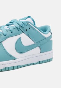 Nike-tennis kengät, joissa on valkoinen ja turkoosi nahkapinta, teksturoitu kumipohja, pyöreä kärki ja litteät nauhat.