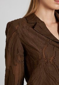 Blusa marrone con un design paisley testurizzato, dotata di colletto e tessuto trasparente. Le cuciture distintive aggiungono profondità e un aspetto stratificato.