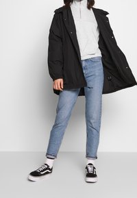 Chaqueta acolchada negra con un dobladillo ajustable, sudadera gris con cremallera debajo, jeans de color azul claro y zapatillas negras con acentos blancos.
