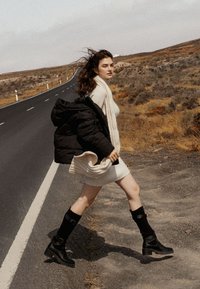 Mujer con un vestido de punto crema, chaqueta de plumas negra y botas altas negras. El conjunto presenta una tela texturada y un silueta ajustada.