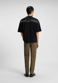 Camiseta negra con el texto "Hilos de Rebelión" en costura de contraste, combinada con pantalones marrones y zapatos negros. Vista posterior.