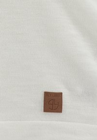 Tessuto bianco lavorato a maglia con una finitura testurizzata, dotato di un'etichetta in pelle marrone con logo in rilievo e la scritta "KEY LARGE" nell'angolo inferiore.