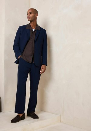 Mann steht auf einer beigen Stufe, trägt einen marineblauen Anzug, ein dunkelbraunes Hemd und dunkelbraune Loafers und schaut nach links vor eine hell strukturierte Wand.