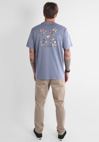 T-shirt en coton bleu clair avec un motif floral dans le dos. Porté avec un pantalon beige. Il a des manches courtes et une coupe décontractée.