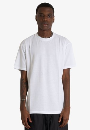 Uomo nero con capelli corti che indossa una maglietta bianca semplice e oversize, pantaloni neri, una collana a catena, orecchini a cerchio e un braccialetto, in piedi rivolto in avanti.
