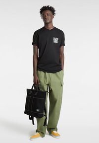 Sort bomulds T-shirt med grafisk print, grønne cargo bukser og sort canvas tote bag med hvid syning. Gule sko fuldender outfittet.