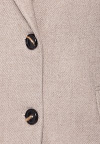 Beige,texturerad tyg med en synlig svart knapp som har ett subtilt mönster. Nära vy som betonar material och knappdetalj.