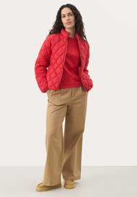 Red quilted jacket with a zip front, paired with a red top, tan wide-legged pants, and beige slip-on shoes. Smooth and warm appearance.