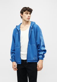 Jauns vīrietis, kurš ir ģērbies zilā rāvējslēdzēja džemperī ar svītrām uz piedurknēm, baltā krekliņā un melnās biksēs, skatās uz sāniem.