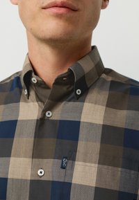 Button-up shirt with a check pattern in brown, navy, and beige shades. Features a collar, contrasting buttons, and a sewn pocket accent.