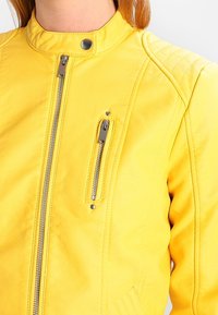 Giubbotto di pelle gialla con colletto rialzato, tasche con zip e dettagli cuciti. Presenta accenti in metallo argentato e una texture liscia.
