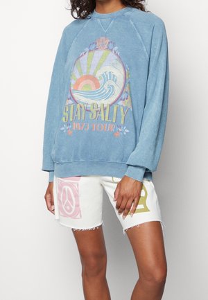 Person iført blå sweatshirt med farverigt "Stay Salty 1973 Tour" bølge- og solgrafik, kombineret med hvide shorts med fredssymbol og bogstaver.