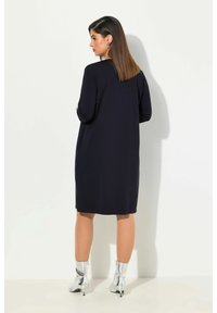 Dunkelblaues Kleid mit langen Ärmeln, knielang, glatte Textur und lockere Passform. Kombiniert mit metallic-silbernen Knöchelstiefeln.