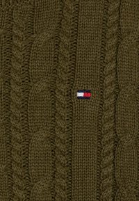 Olivgrüner Kabelstrickstoff mit kleinem rechteckigem, rot-weiß-blauem gesticktem Logo in der Mitte.