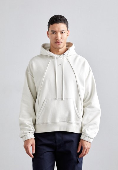 Lys beige pullover hoodie i blød stof, med en frontlomme, snoretræk og ribbede manchetter. Modellen har mørke bukser på.