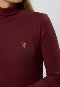 Detailný záber na osobu, ktorá má na sebe ryhovaný bordový rolák s malým vyšitým logom polo hráča na hrudi.