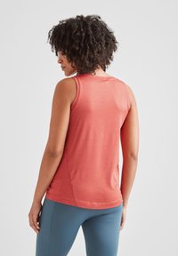 Woman with curly hair wearing sleeveless coral top and blue leggings, facing away against a white background.