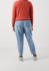 Haut orange à manches longues associé à un jean bleu clair à coupe décontractée. Le jean comporte des poches arrière ; le modèle porte des chaussures de sport gris clair.
