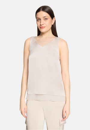 Kvinde med glat mørkt hår iført en ærmeløs beige top og lyse beige bukser med sidelomme mod en ensfarvet baggrund.