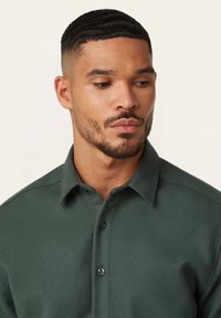 Donkergroene overhemdd met een gestructureerde stof, met een puntige kraag en zwarte knopen, weergegeven op een mannelijke model tegen een lichte achtergrond.